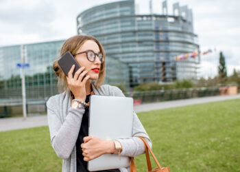 Lifestyle portrait of a young woman standing with laptop and smartphone in front of the European parliament building in Strasbourg, France