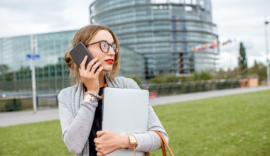 Lifestyle portrait of a young woman standing with laptop and smartphone in front of the European parliament building in Strasbourg, France